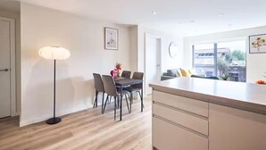 A modern kitchen with a dining area, featuring a wooden floor, a dining table with chairs, a pendant light, and a large window with a view of the outside.