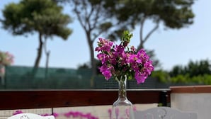 Une table blanche sur laquelle est posé un vase en verre transparent contenant des fleurs roses, le tout placé sur une chaise blanche agrémentée d’un dessous de verre en osier.