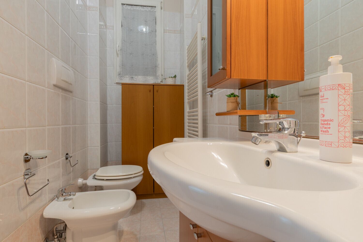 Un bagno con lavandino bianco, un water e un bidet. Sopra il lavandino c'è un mobiletto in legno e uno specchio con ripiano, dove è posizionata una pianta.