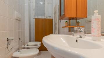 Un bagno con lavandino bianco, un water e un bidet. Sopra il lavandino c'è un mobiletto in legno e uno specchio con ripiano, dove è posizionata una pianta.