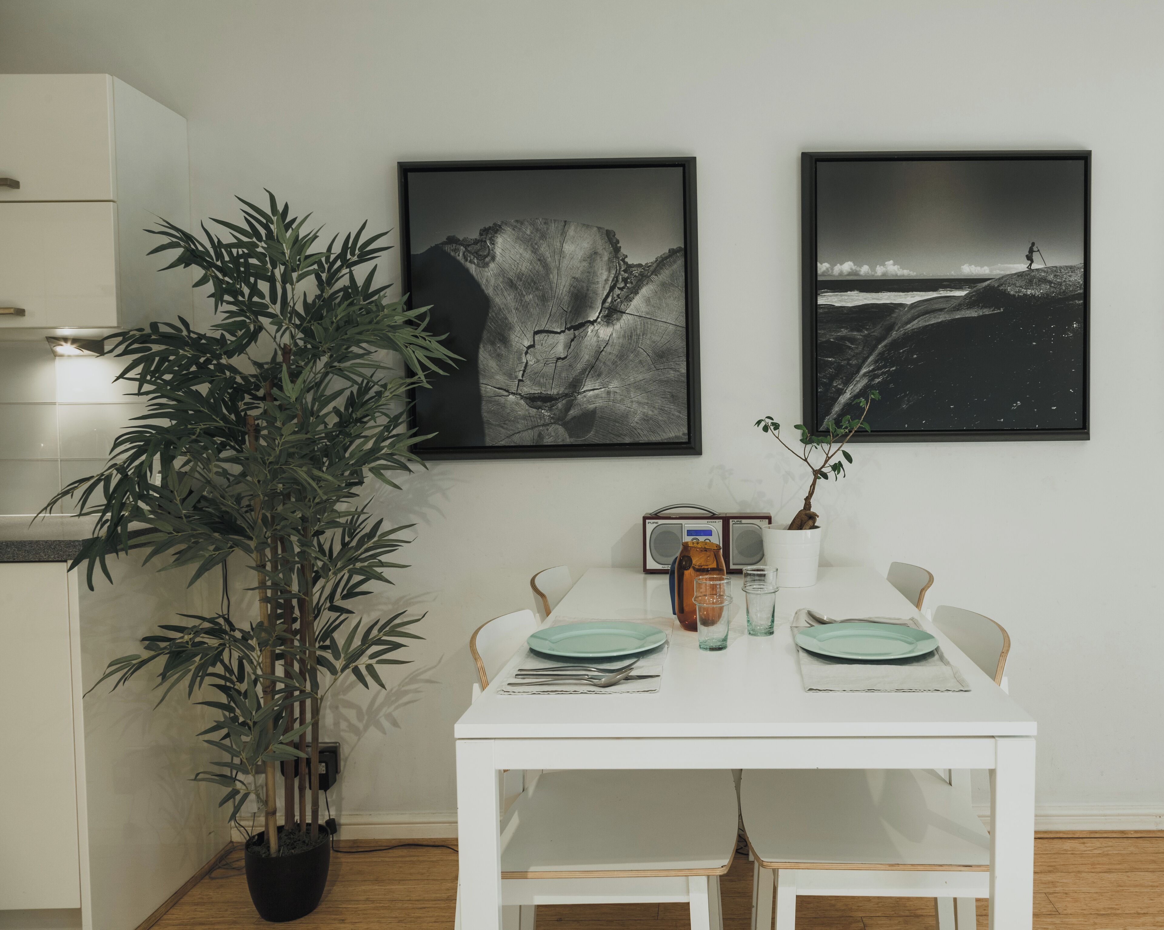 A dining area with a white table set for two, two framed black and white photographs on the wall, a radio, and a potted plant.