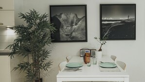 A dining area with a white table set for two, two framed black and white photographs on the wall, a radio, and a potted plant.