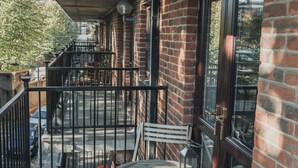 A balcony with a wooden table and chairs, a metal railing, and a brick building in the background.