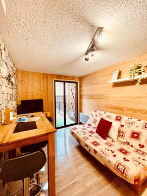 A cozy room with a wooden floor, a sofa with patterned cushions, a desk with a computer, and a sliding glass door leading outside. - Studio/ terrace "le Plan sec", 3 pers Aussois (Aussois)