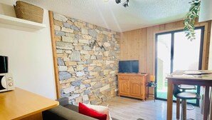 Un espace de vie avec un mur en pierre, un plafond en bois, un téléviseur, un coin cuisine avec un tabouret de bar et une fenêtre.