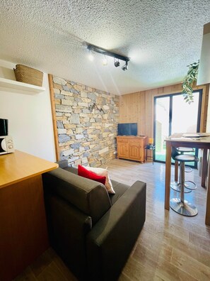 A living area with a stone wall, a wooden ceiling, a television, a kitchen area with a bar stool, and a window. - Studio/ terrace "le Plan sec", 3 pers Aussois (Aussois)
