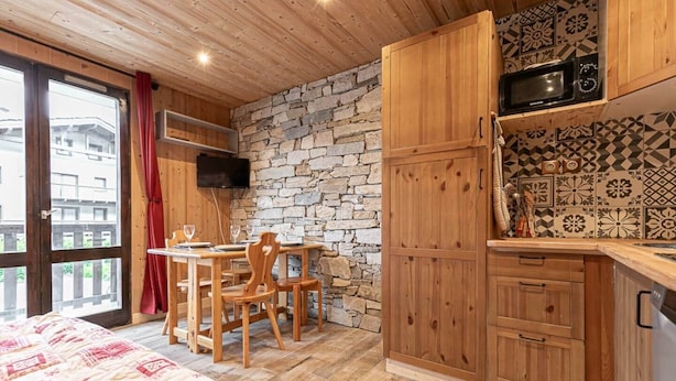 A cozy room with a bed, a dining table with chairs, a stone wall, wooden cabinets, and a TV mounted on the wall.
