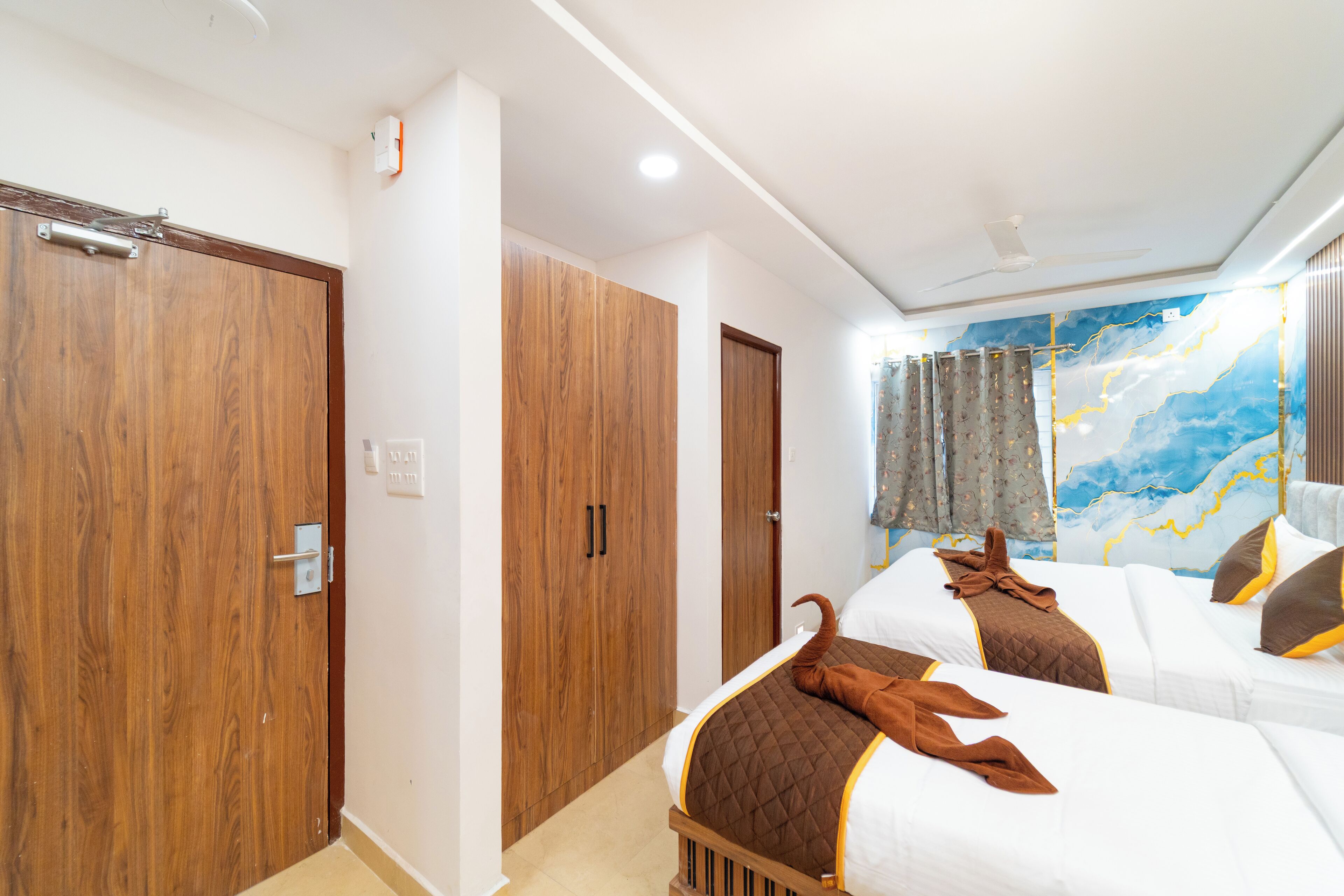 A hotel room with two beds, wooden wardrobes, a ceiling fan, and a large abstract painting on the wall.