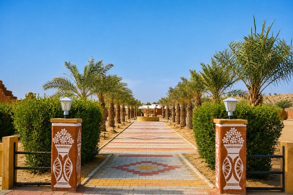 A pathway leading to a building with a patterned tile floor, flanked by palm trees and decorative pillars with floral designs. - barna dersert camp jaisalmer (Jaisalmer)