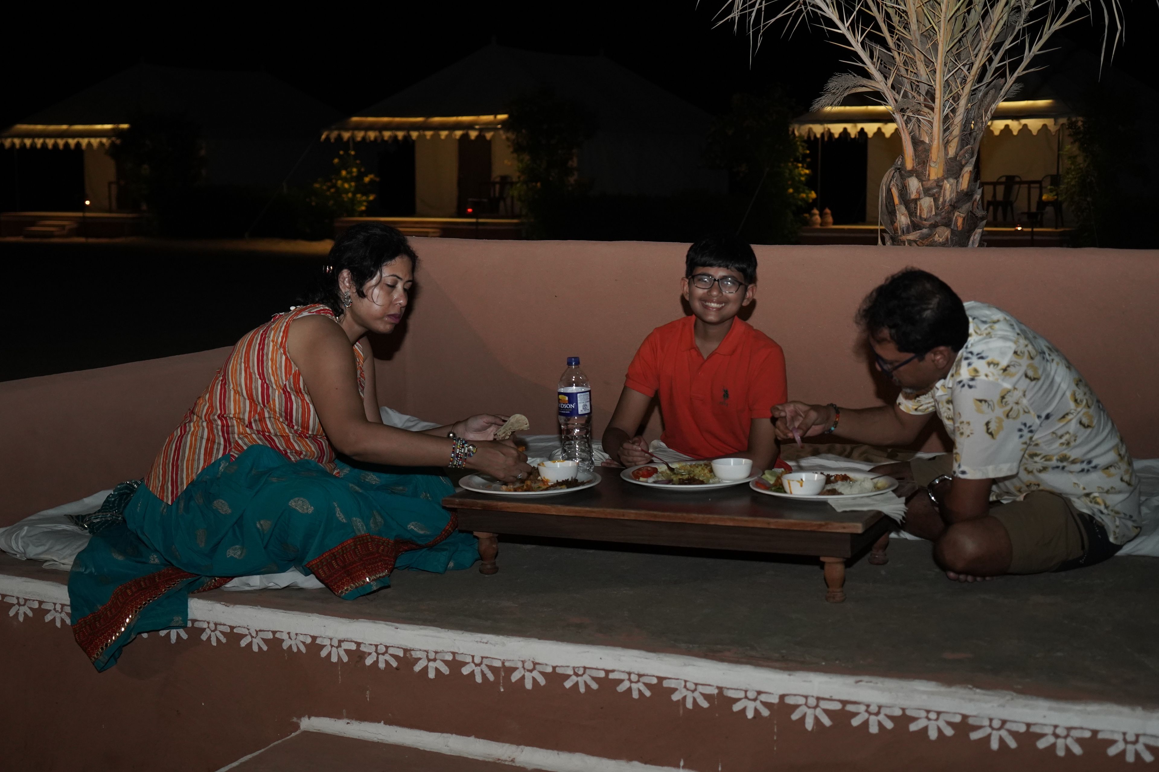 Tres personas sentadas en un banco al aire libre por la noche, comiendo de platos.