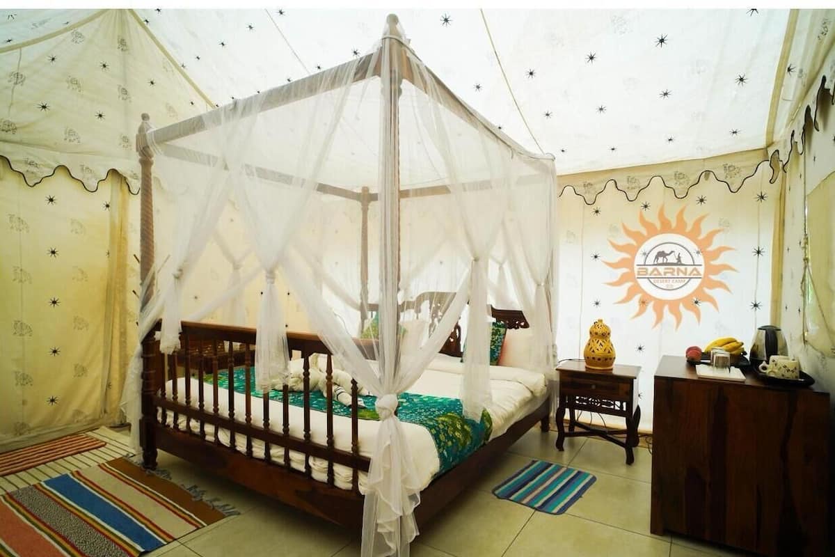 A room with a four-poster bed, a small wooden side table, a wall-mounted radiator, and a decorative wall with a sun emblem.