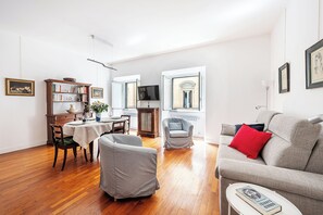A living room with a grey sofa, a dining table with chairs, a bookshelf, and a television.