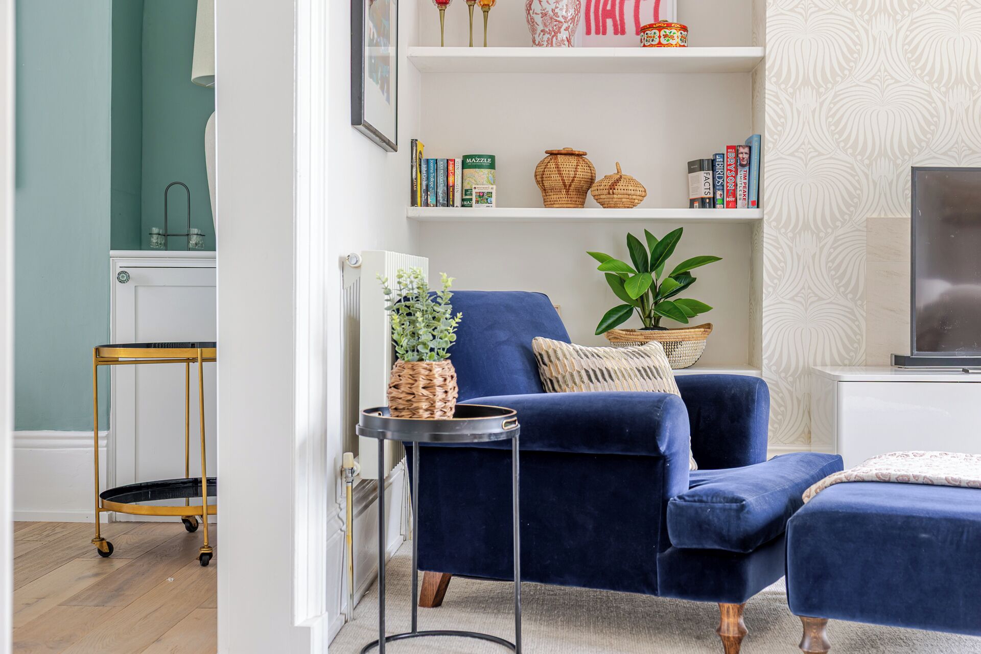 Uma sala de estar com uma poltrona azul, uma mesinha lateral com uma planta, uma estante com livros e objetos decorativos, e uma televisão sobre um móvel.