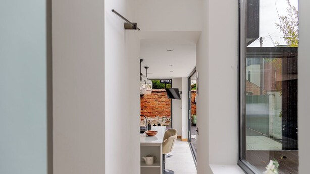 A narrow hallway leading to a kitchen with a brick wall, modern furniture, and a large window.