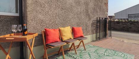 Une terrasse avec une table en bois, deux chaises, une bouteille de vin et un tapis à motifs verts.