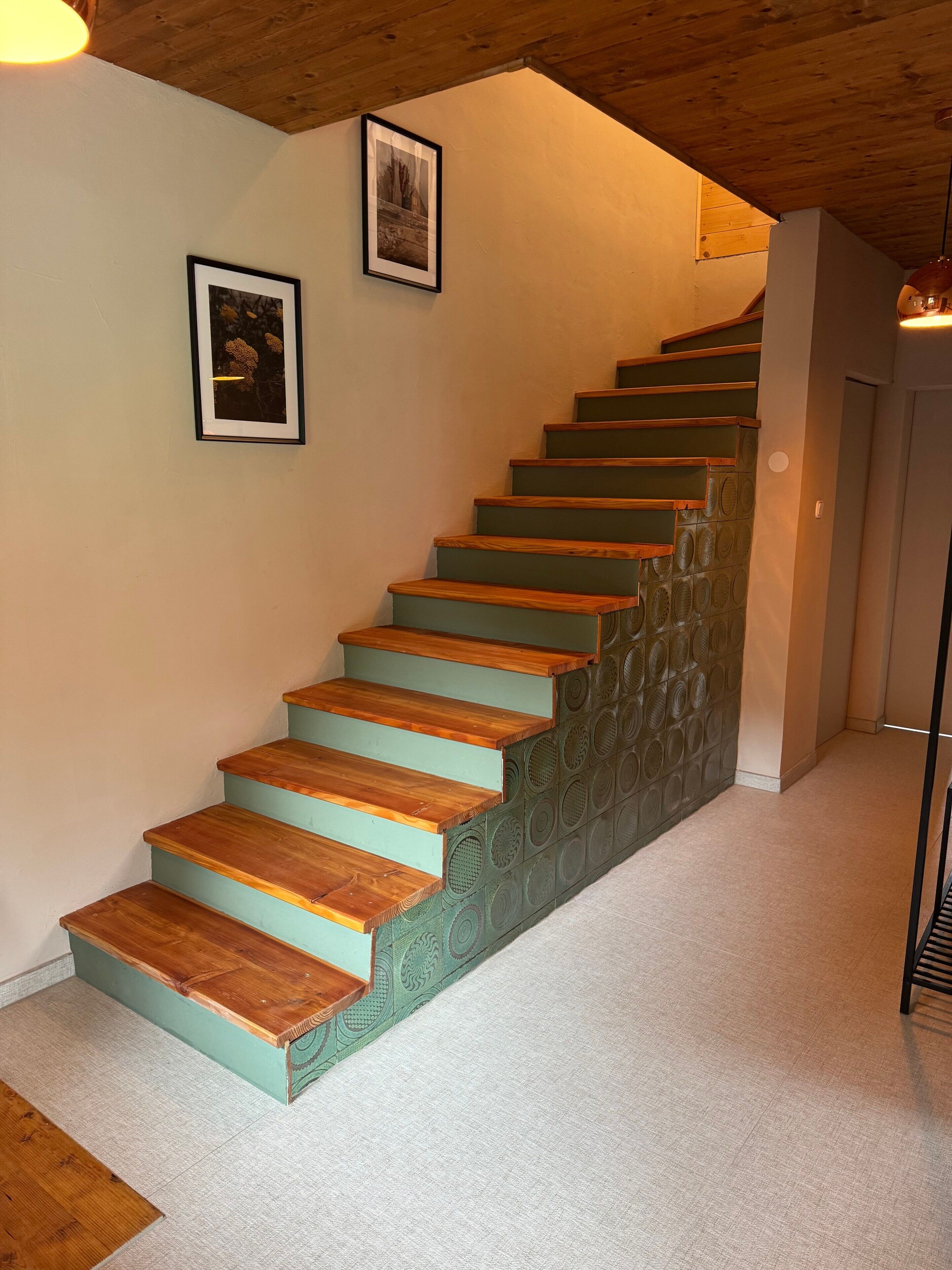 Een trap met afwisselend houten en groene treden, twee ingelijste foto's aan de muur en een houten plafond.