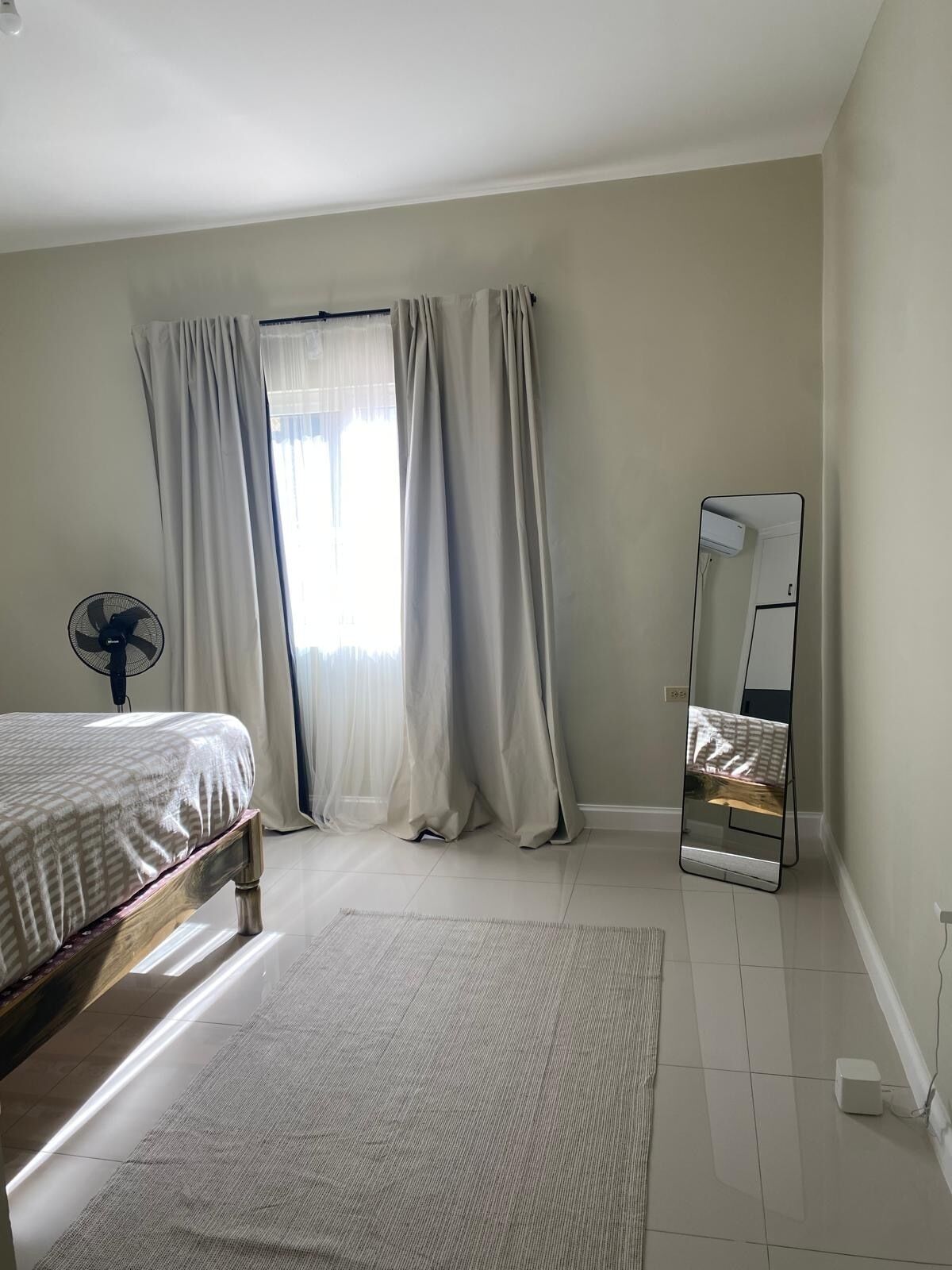 Een slaapkamer met een bed, een ventilator, een spiegel en een raam met gordijnen.