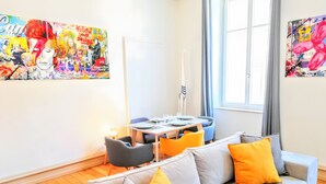 Una sala de estar moderna con un sofá gris, cojines amarillos, una mesa de comedor para cuatro personas y dos grandes cuadros abstractos en las paredes.