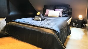 A bedroom with a sloped ceiling, a skylight, a bed with a black mattress and pillows, a bedside table with a lamp, and a box labeled '20 Liters' on the floor.
