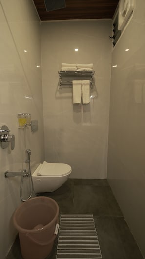 A bathroom with a toilet, a shower, a towel rack, a bucket, and a hand dryer.