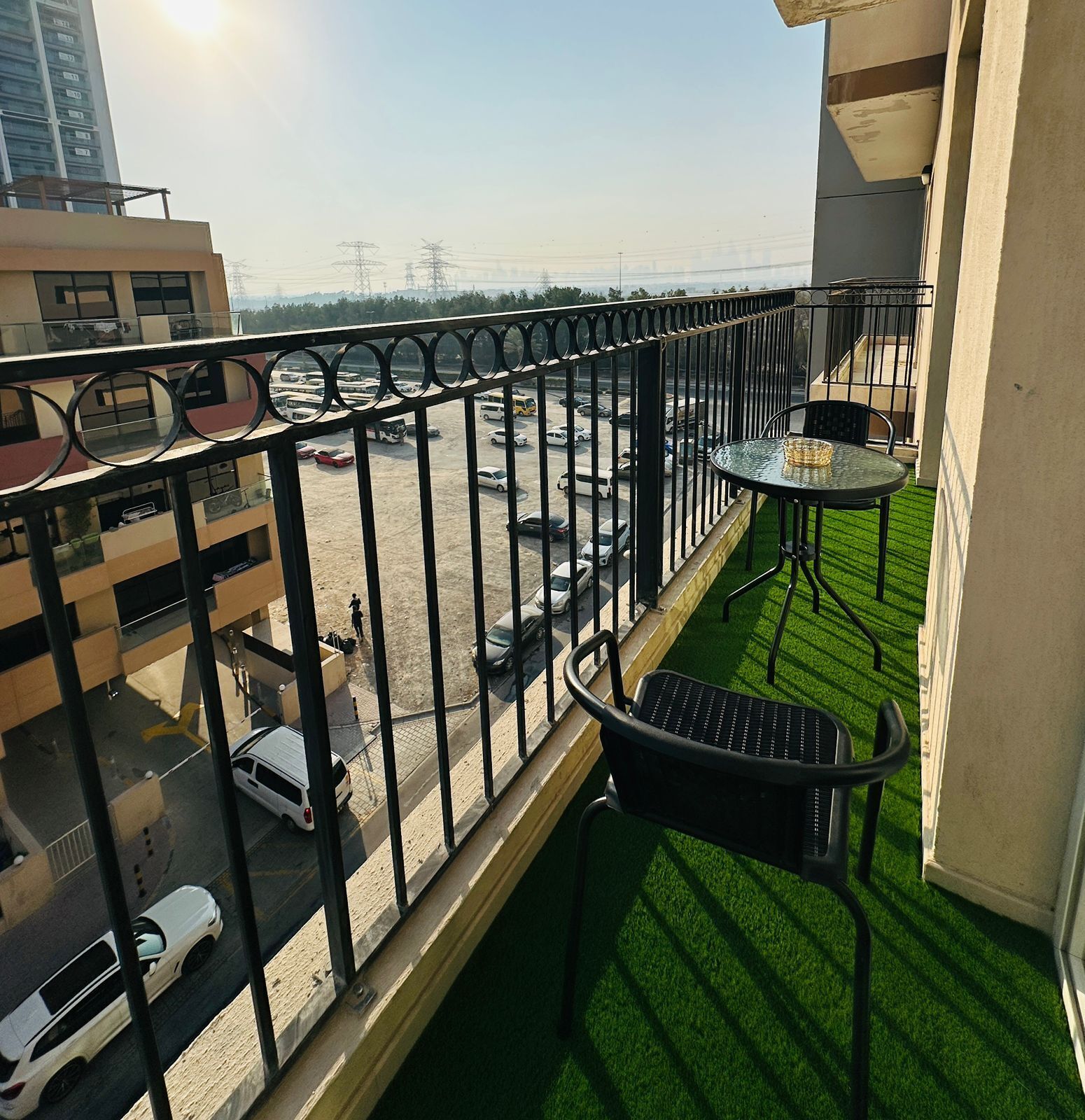 A balcony with a small table and chairs, a view of a parking lot with various vehicles, and a building in the background.