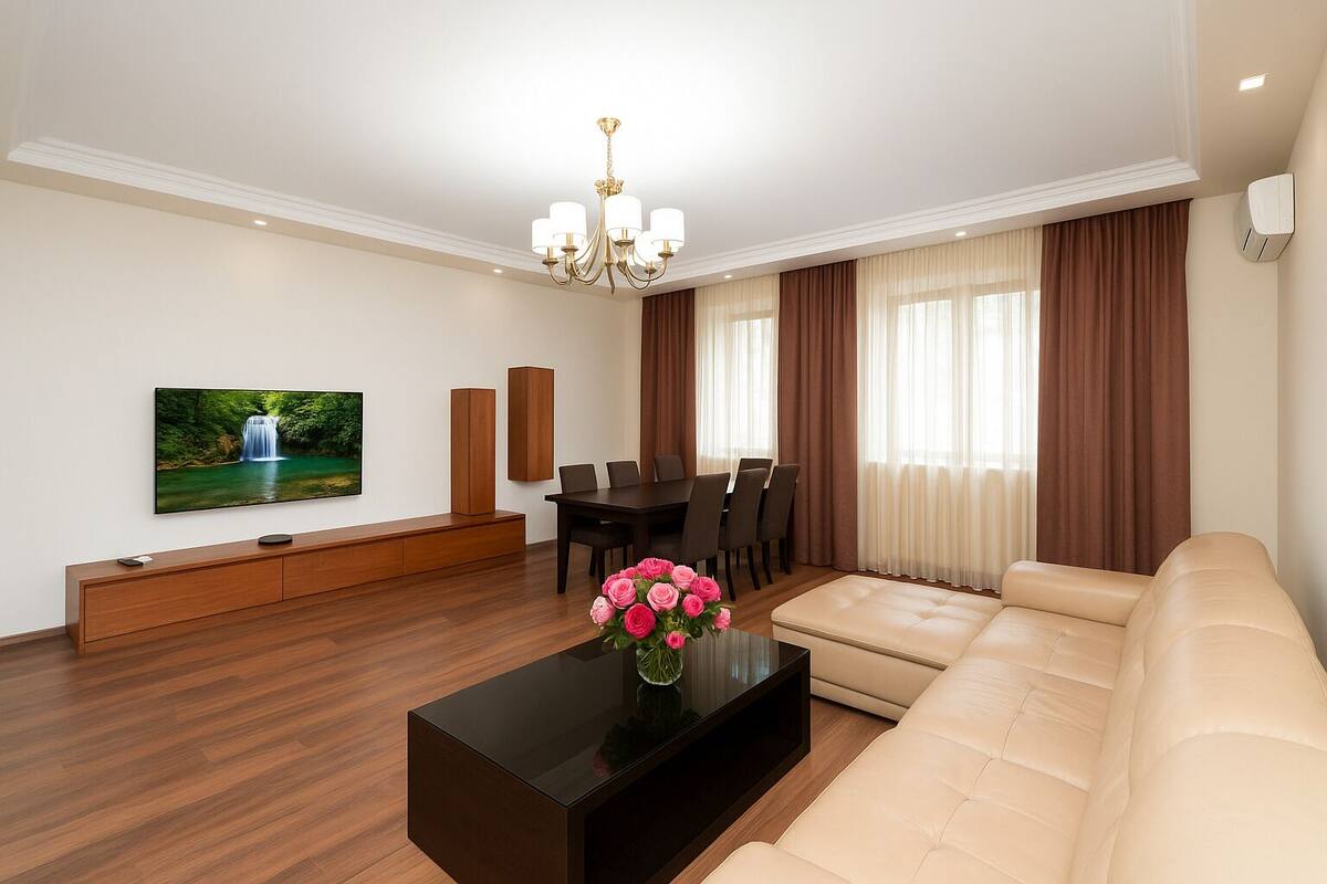 A modern living room with a flat-screen TV, a wooden entertainment unit, a glass coffee table with flowers, a beige sofa, and a dining area with a table and chairs.