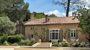A two-story house with a tiled roof, a front entrance with a set of steps, and a small garden area with various plants and a seating area with outdoor furniture. - Escape to Elegance -Wonderful Mas Provencal in the heart of the Alpilles (Saint Etienne du Grés)