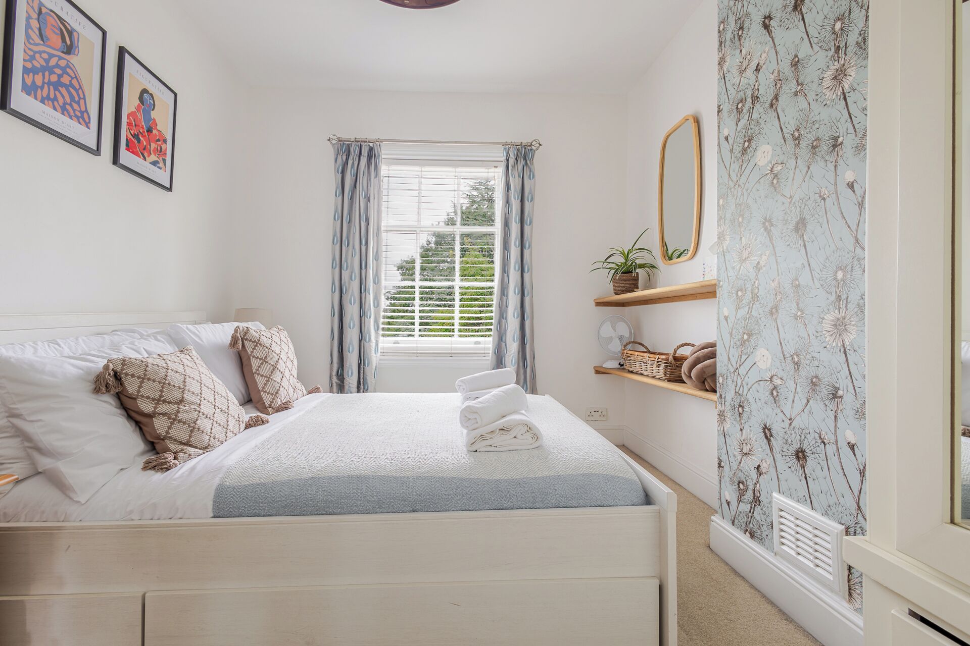 A neatly made bed with a white headboard, a bedside table with a lamp, a window with curtains, and a wall with framed artwork.