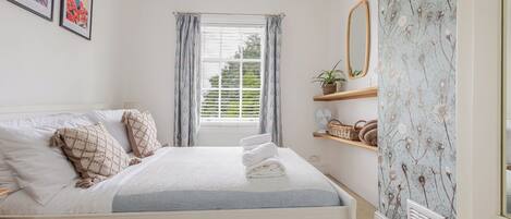 A neatly made bed with a white headboard, a bedside table with a lamp, a window with curtains, and a wall with framed artwork.