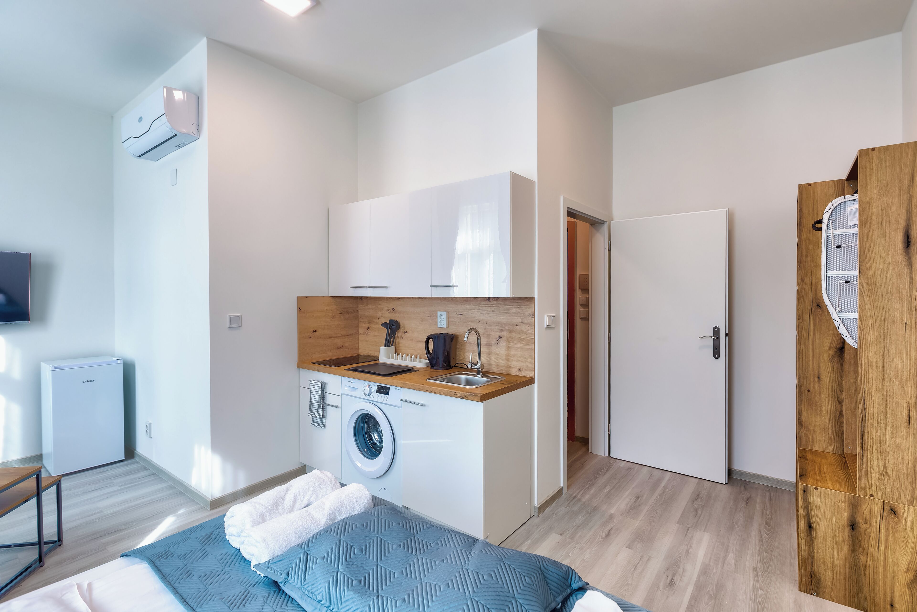 Een compacte, moderne kamer met een wasmachine, een klein kitchenette, een bed met een blauwe sprei en een houten deur.