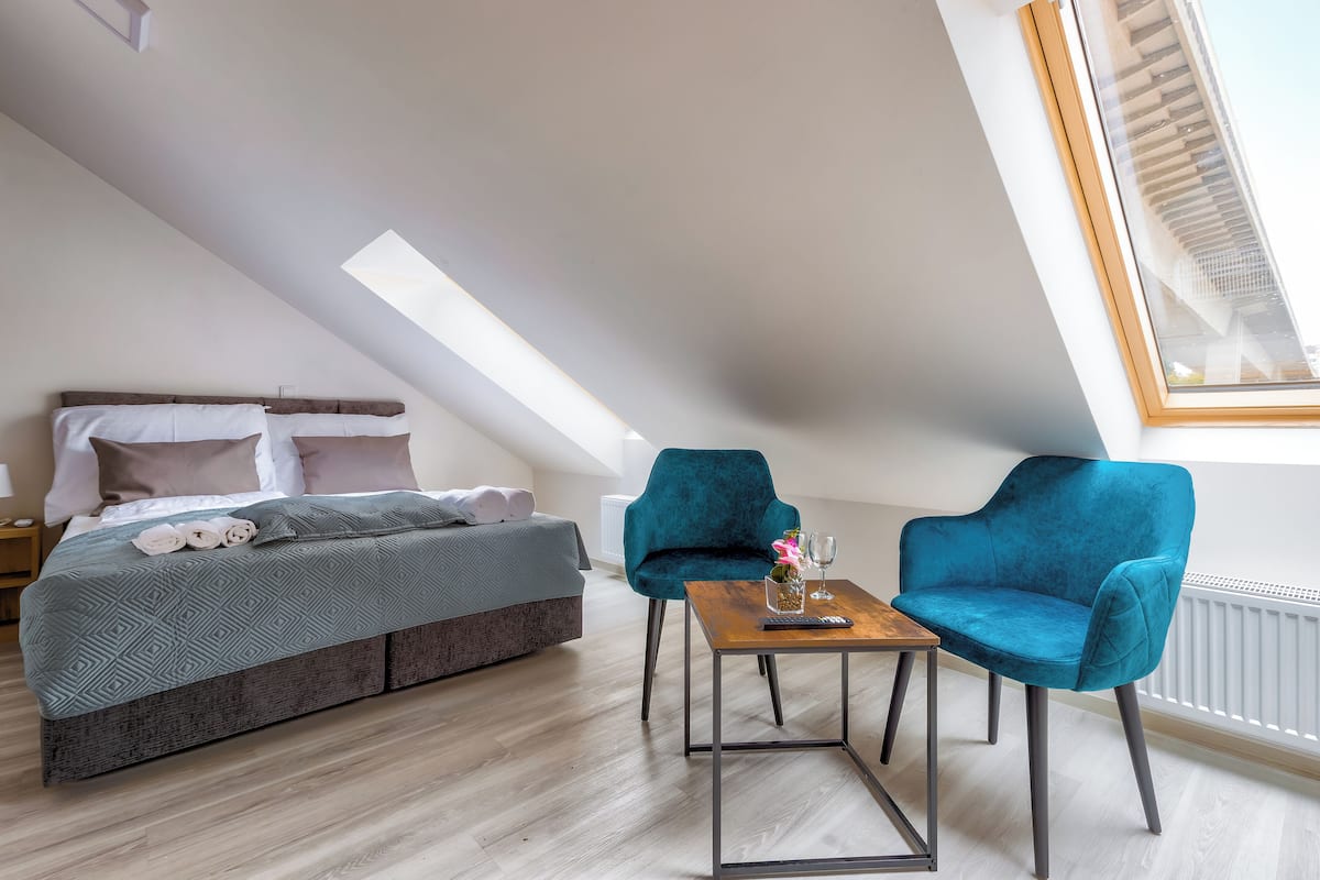 A modern bedroom with a loft-style bed, a small table with a vase and remote control, and two teal armchairs.