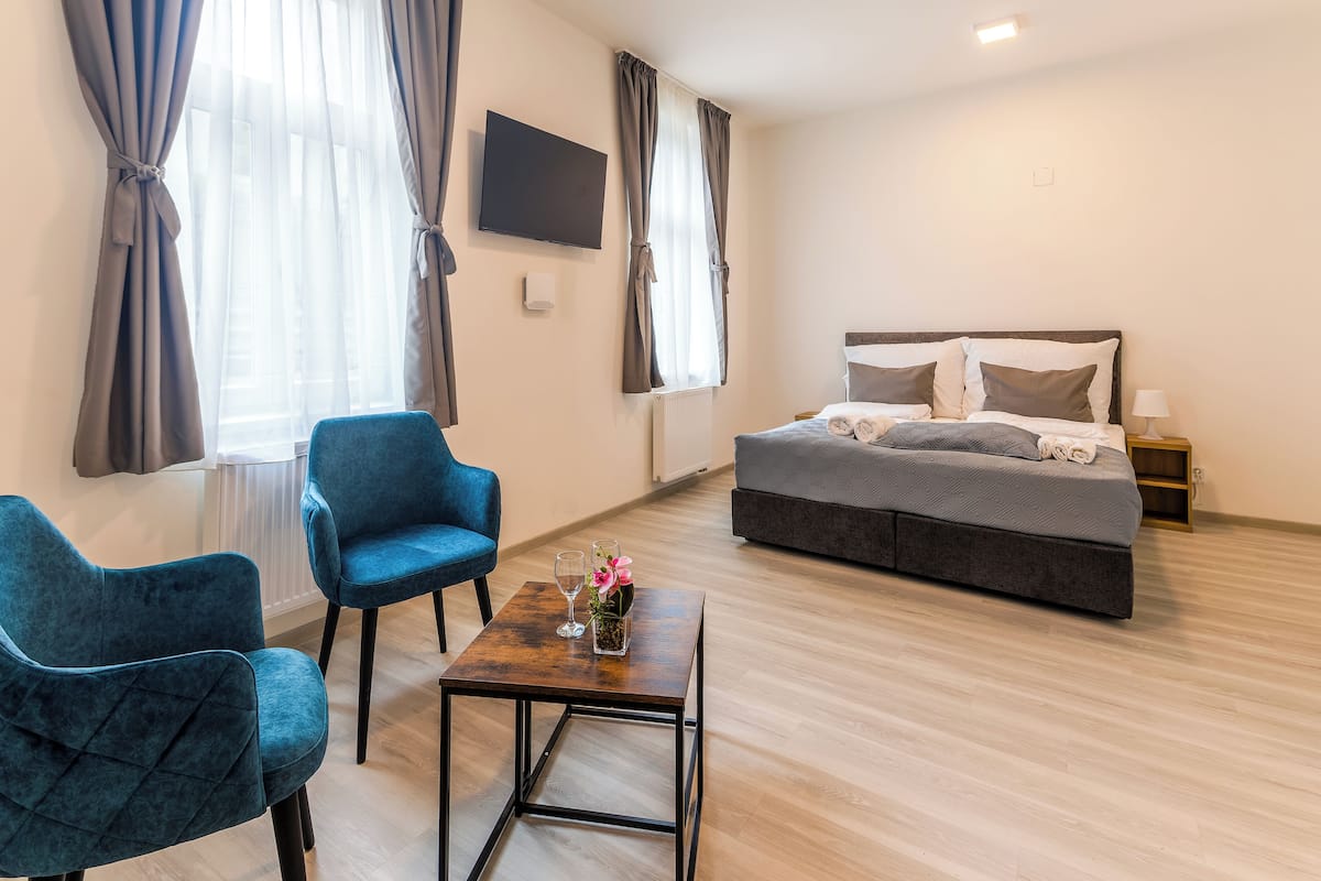 A hotel room with a bed, two blue armchairs, a small table with a vase of flowers, and a TV on the wall.