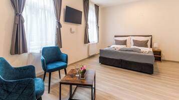 Une chambre d’hôtel avec un lit, deux fauteuils bleus, une petite table sur laquelle est posé un vase de fleurs et un téléviseur fixé au mur.