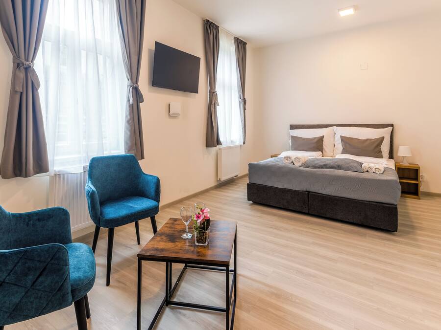 Une chambre d’hôtel avec un lit, deux fauteuils bleus, une petite table sur laquelle est posé un vase de fleurs et un téléviseur fixé au mur.