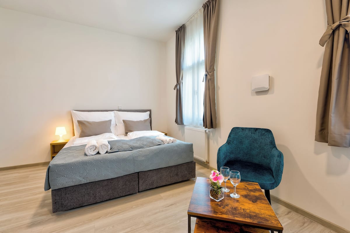 A hotel room with a bed, a nightstand, a table with a vase and glasses, and a chair.