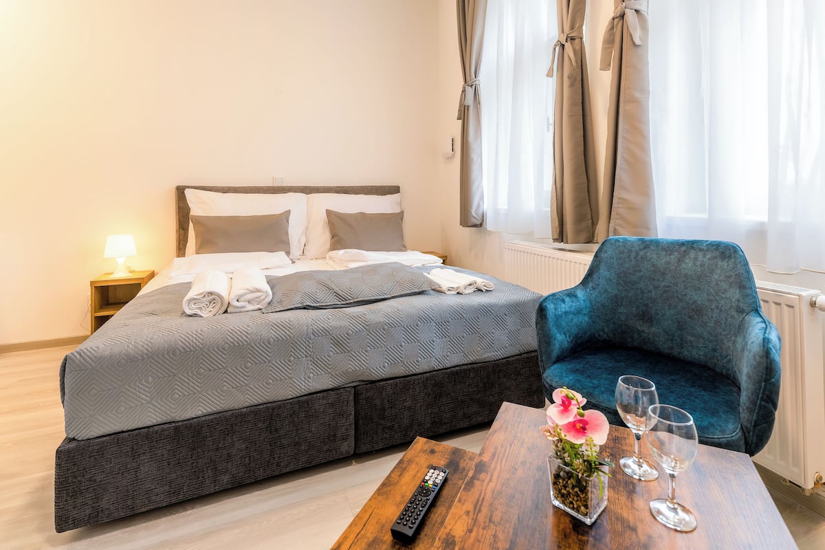A hotel room with a bed, a blue armchair, a wooden coffee table with a remote control, a vase with flowers, and two wine glasses.