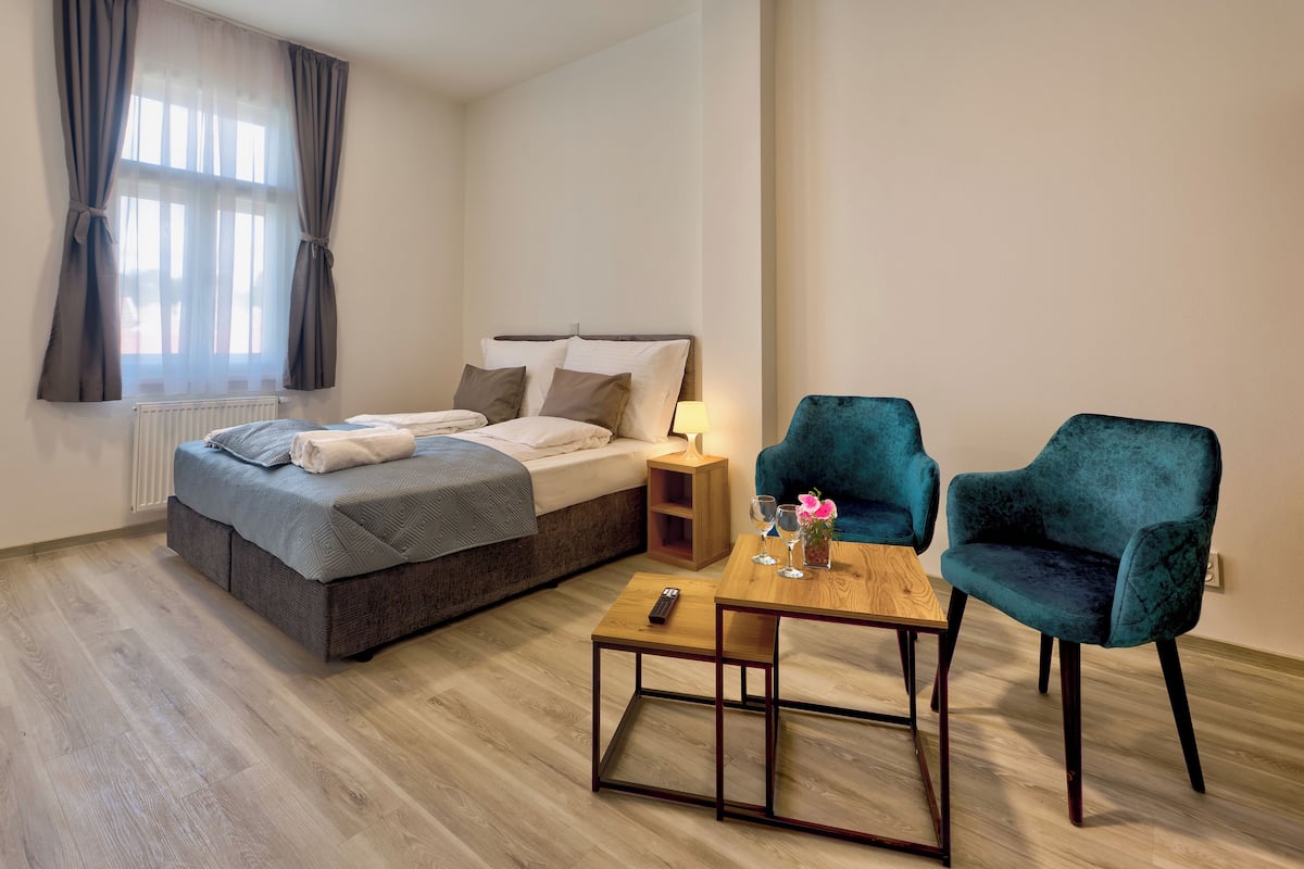 A bedroom with a bed, bedside table, two chairs, and a small table with a remote control.