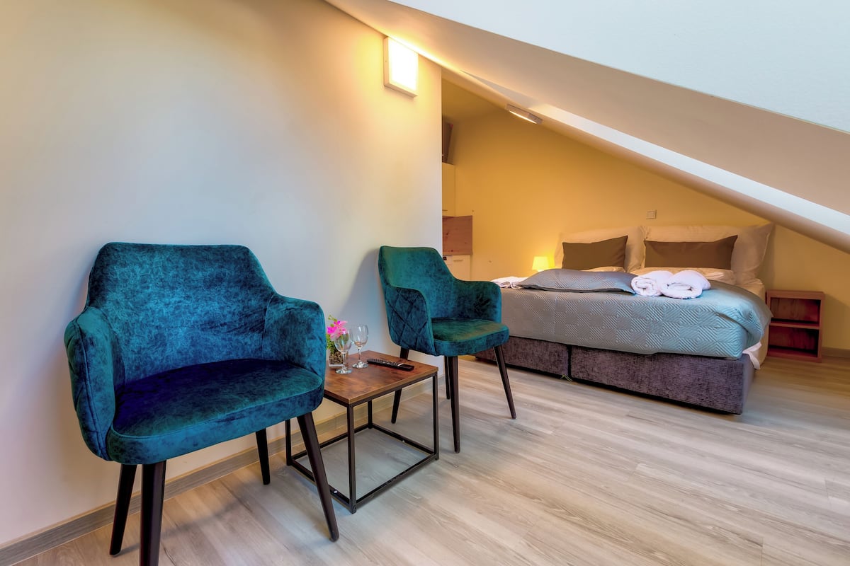 A modern bedroom with a loft-style bed, two teal armchairs, a small table with a vase, and a nightstand.