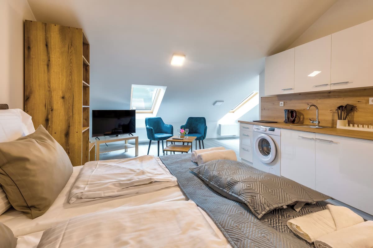 A compact bedroom with a bed, a desk, and a chair. There is a washing machine and a small table with a vase.