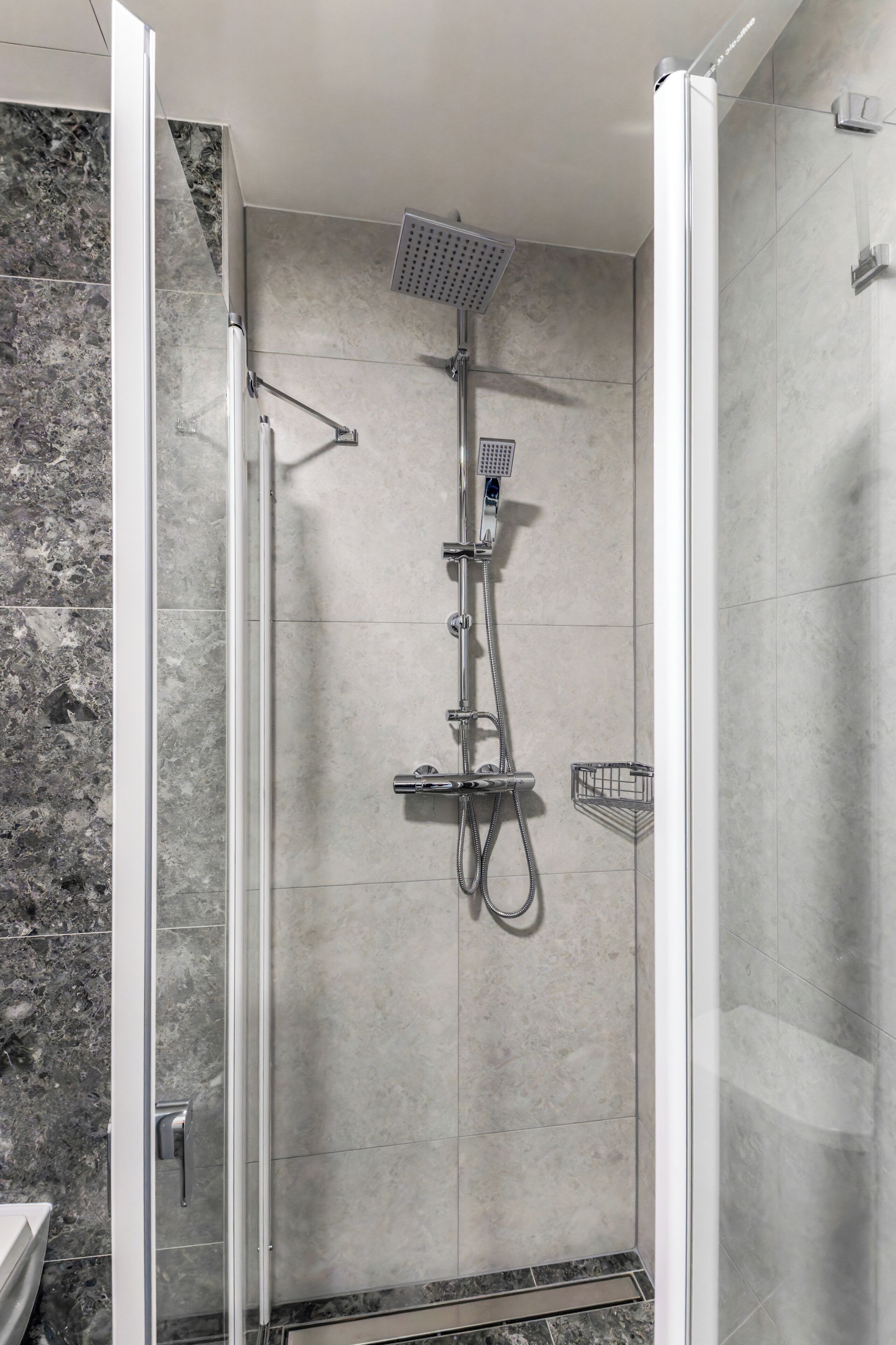 A walk-in shower with a glass door, a handheld showerhead, a wall-mounted showerhead, and a small shelf.