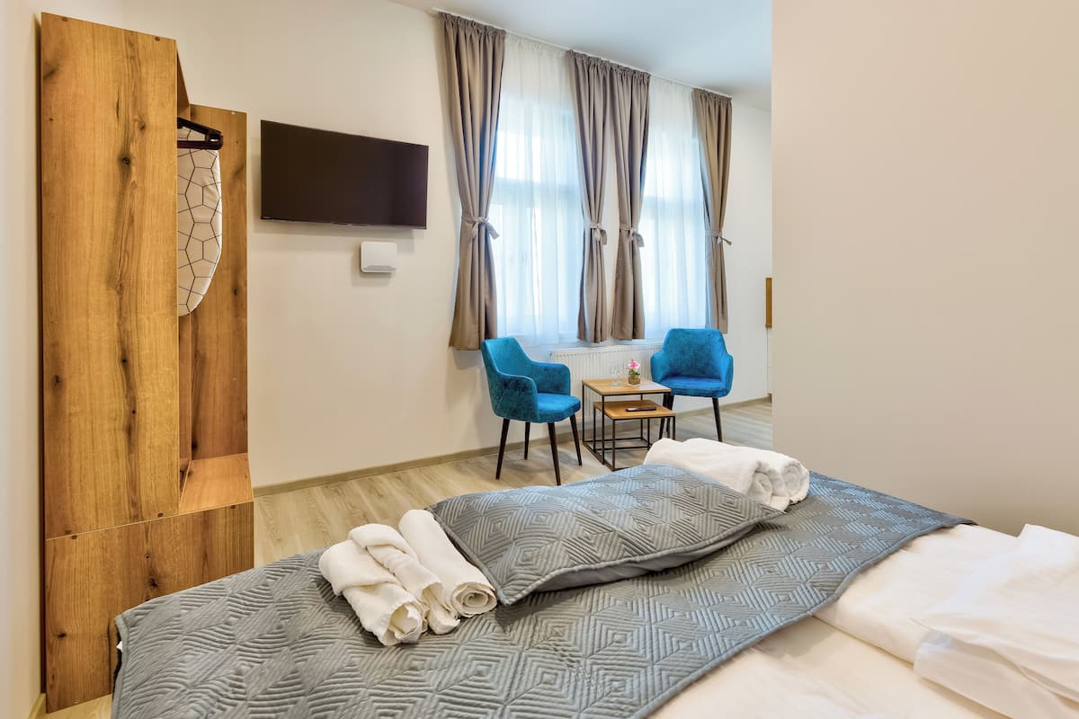 A hotel room with a bed, two blue chairs, a small table, and a TV on the wall.