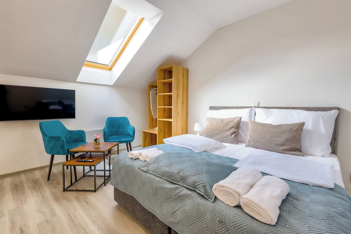 A modern hotel room with a skylight, a bed with white linens, a wooden nightstand, a blue chair, and a TV mounted on the wall.