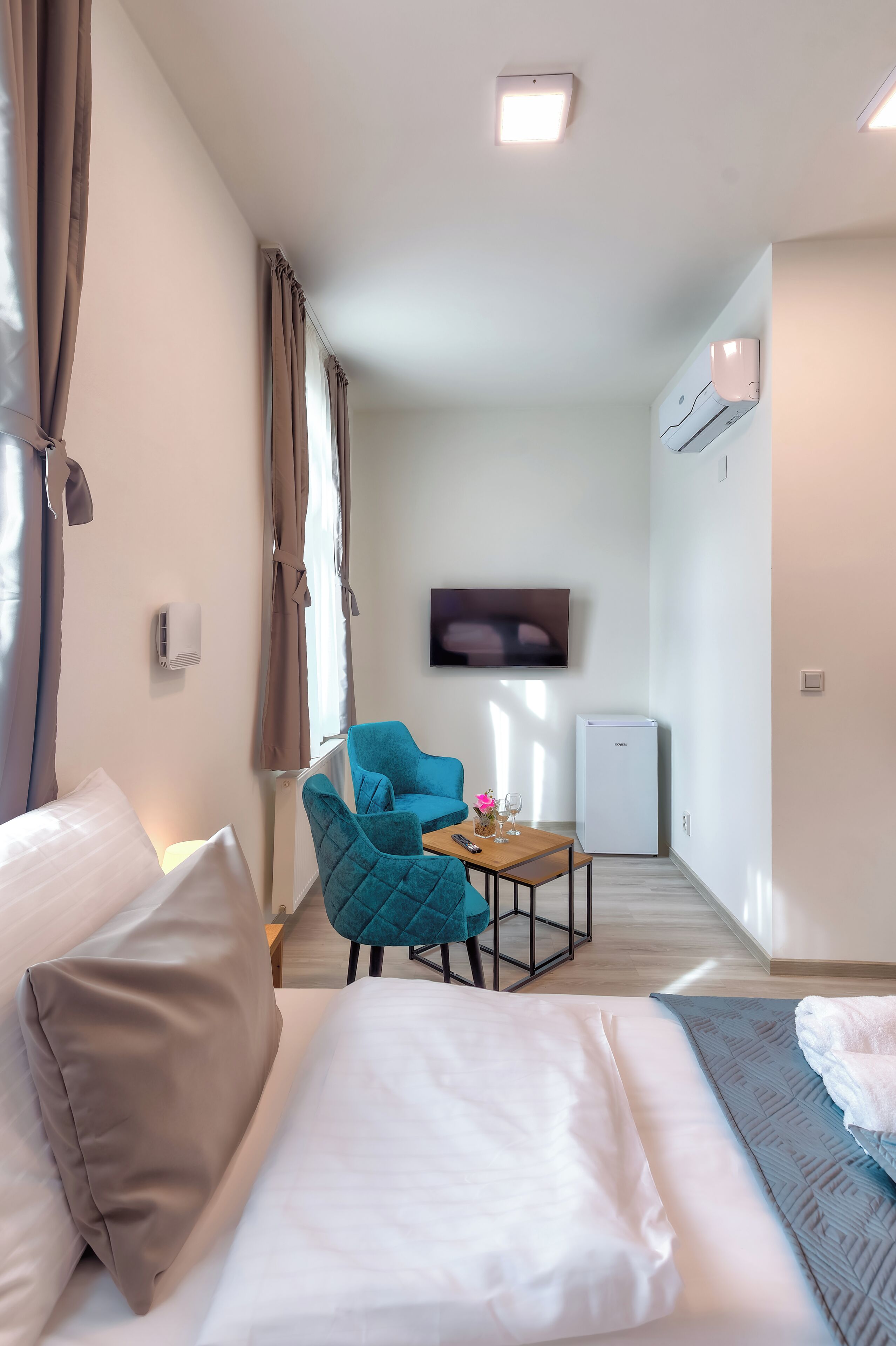 Ein modernes Hotelzimmer mit Sofa, einem kleinen Tisch, einem blauen Sessel, einem an der Wand befestigten Flachbildfernseher und einem Minikühlschrank.