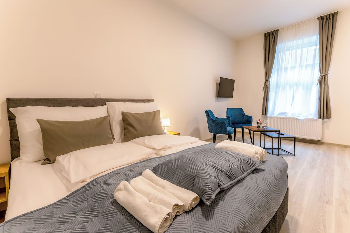 A neatly made bed with a grey patterned blanket and two rolled towels, a blue chair, a small table with a glass, and a TV on the wall.