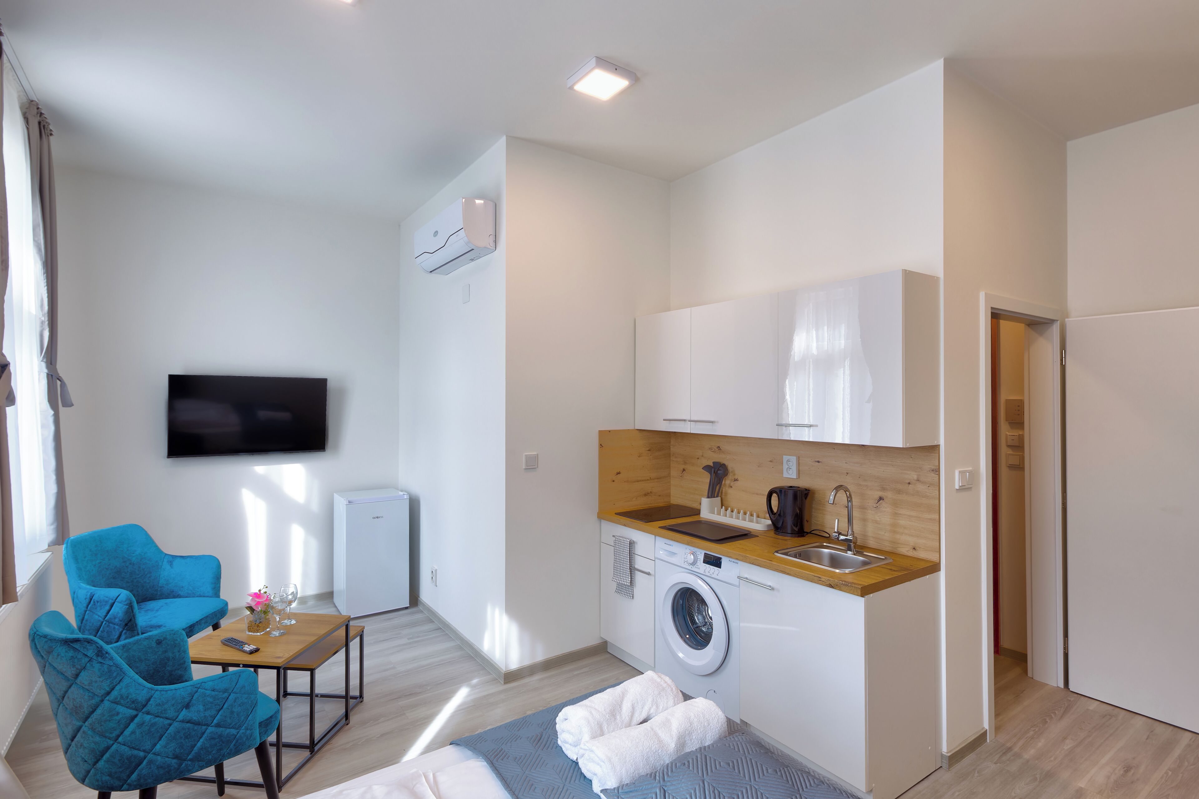 Un espace de vie compact comprenant un coin cuisine, un lave-linge, un téléviseur à écran plat et un coin salon avec deux fauteuils.