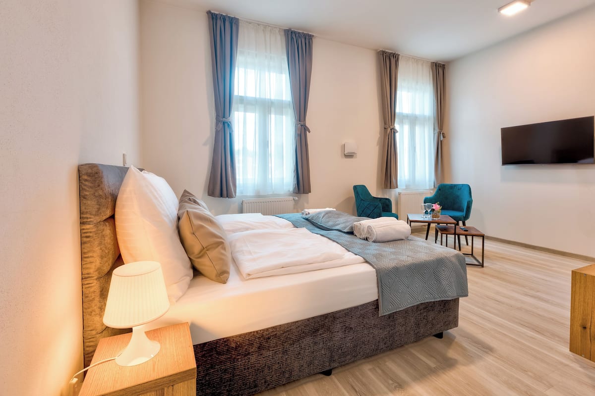 A neatly made bed with pillows and a blanket, a bedside lamp, a chair, a small table with a vase, and a TV mounted on the wall.