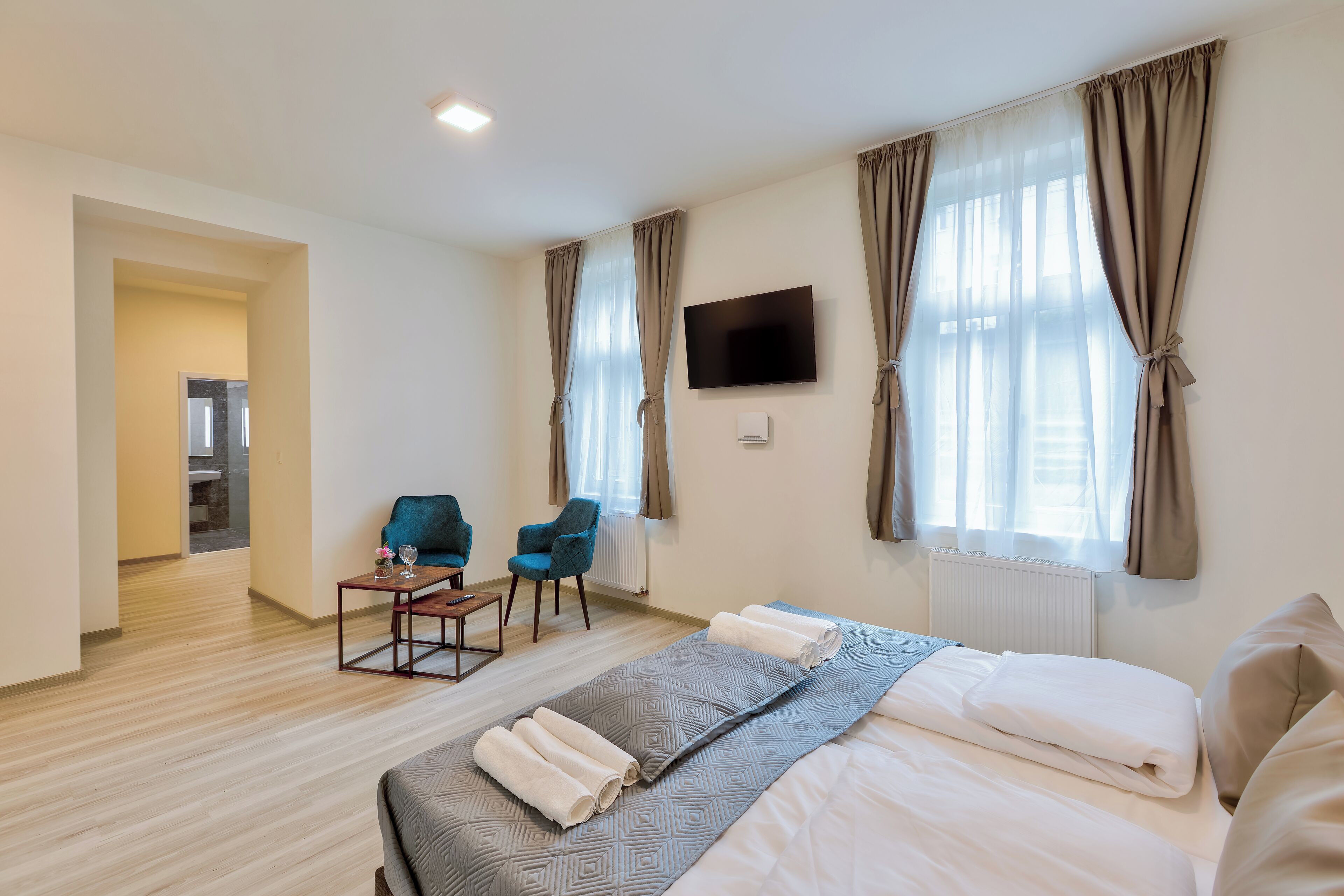 A hotel room with a bed, a TV, a seating area with chairs, and a view of the outside through large windows.