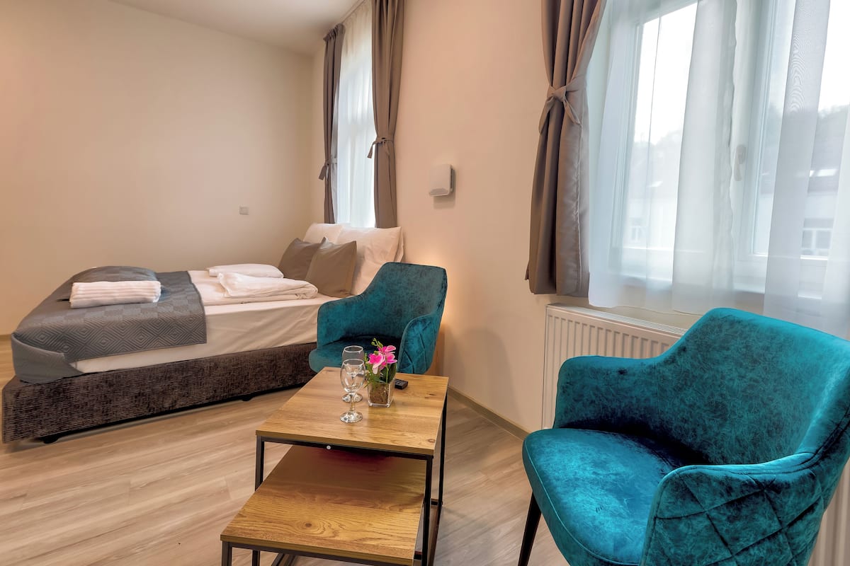 A bedroom with a bed, a wooden table, two teal chairs, and a window with curtains.