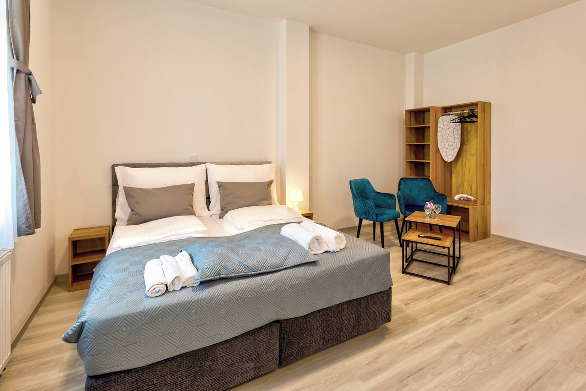 A neatly made bed with pillows and folded towels, a small wooden nightstand, a blue chair, a wooden shelf with a towel, and a small table with a glass and remote control.