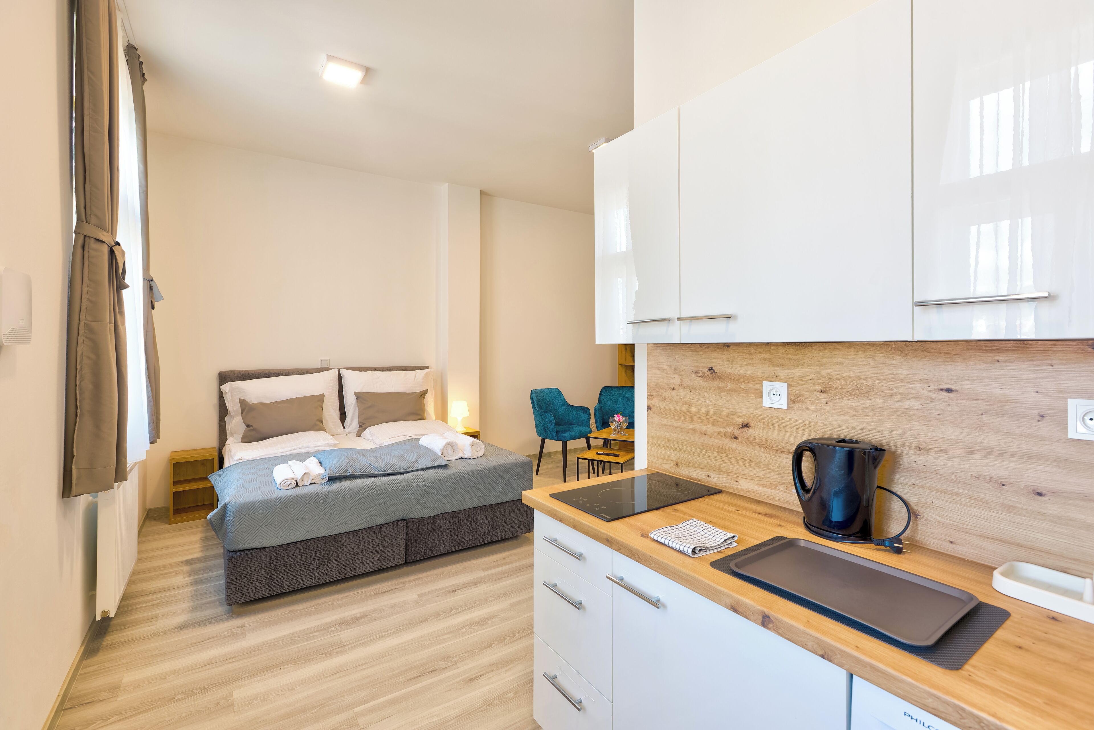 Habitación de hotel moderna con cama, kitchenette, zona de comedor y vistas al exterior.